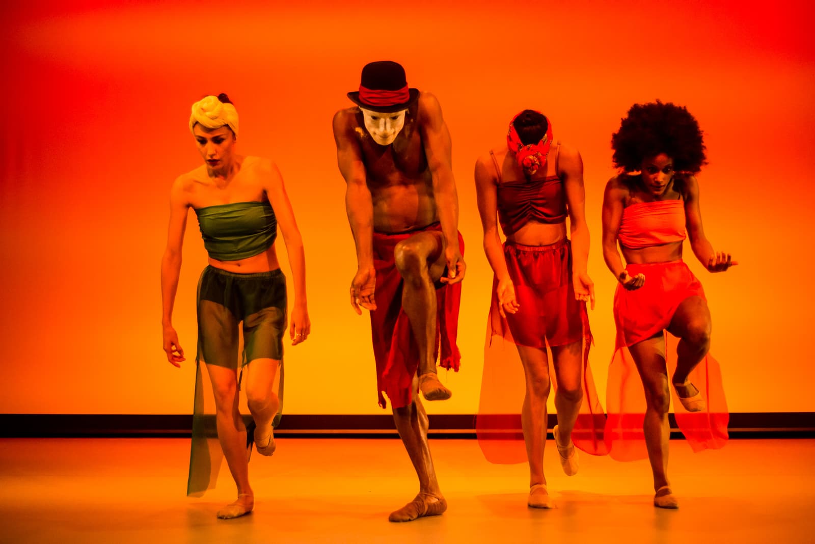 Four dancers performing in a row on a stage with a bright orange background. From left to right: a dancer in green and dark top and skirt, a dancer in a black hat and red skirt with white face paint, a dancer in a red top and red sheer skirt, and a dancer in a red top and red sheer skirt. They are all in motion, captured mid-step with their limbs bent and angled.