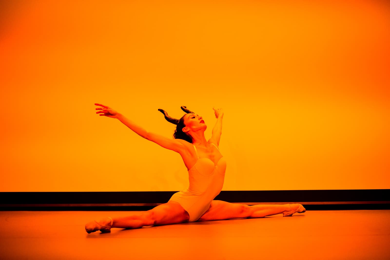 A dancer in a pale leotard and headpiece with curved horns performs a front split on a stage floor against a solid, vibrant orange-lit background, arms extended upwards and outwards, gaze directed towards the upper left corner of the frame.