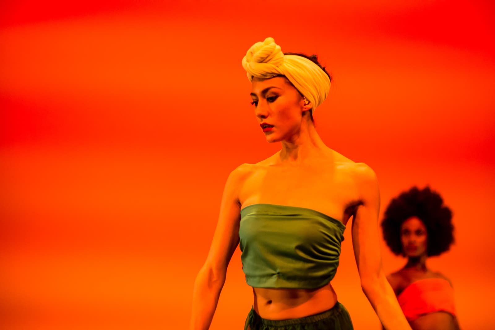 A dancer in the foreground wears a plain green tube top and a light-coloured headwrap, looking down and to the side against a vibrant, solid orange background. Behind them, slightly out of focus, is another person with dark voluminous hair wearing an orange garment.