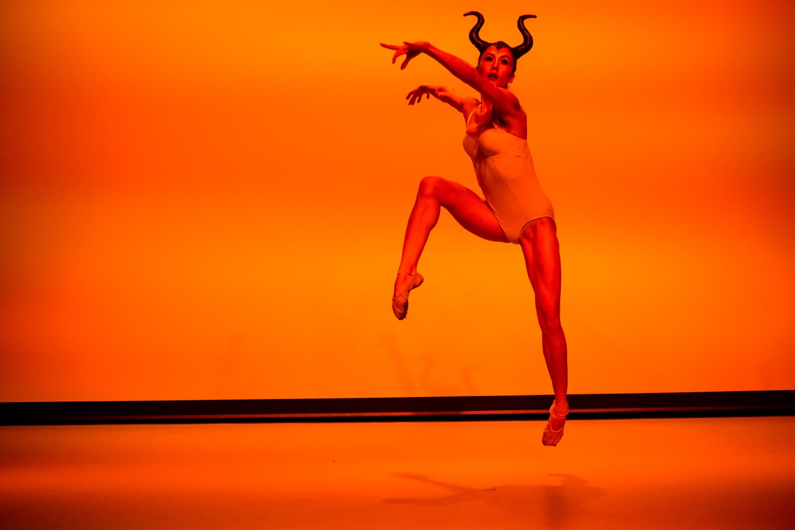 A dancer captured mid-leap against a solid, intense orange background. The dancer is wearing a simple white leotard and an ornamental headpiece with long, curved horns. One leg is bent and raised, while the other is extended downward, with ballet slippers on both feet. The lighting is uniform and warm, casting a slight shadow on the floor beneath the floating figure.