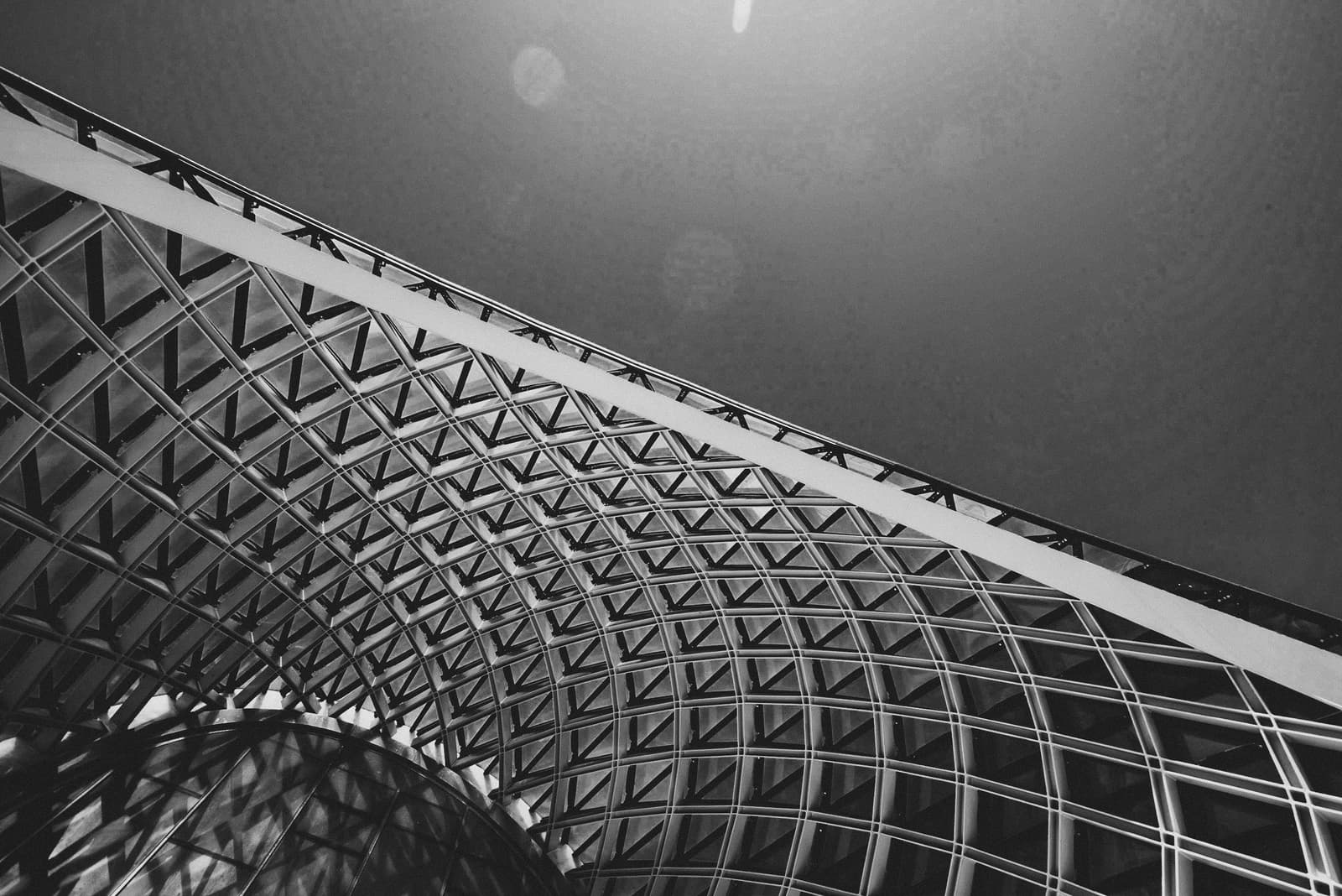 A low-angle, black-and-white architectural shot focusing on a curved, repeating steel diagrid structure against a bright sky. The frame features a solid, curved structural support sweeping across the center, intersecting with a complex network of geometric metal panels and glass segments.