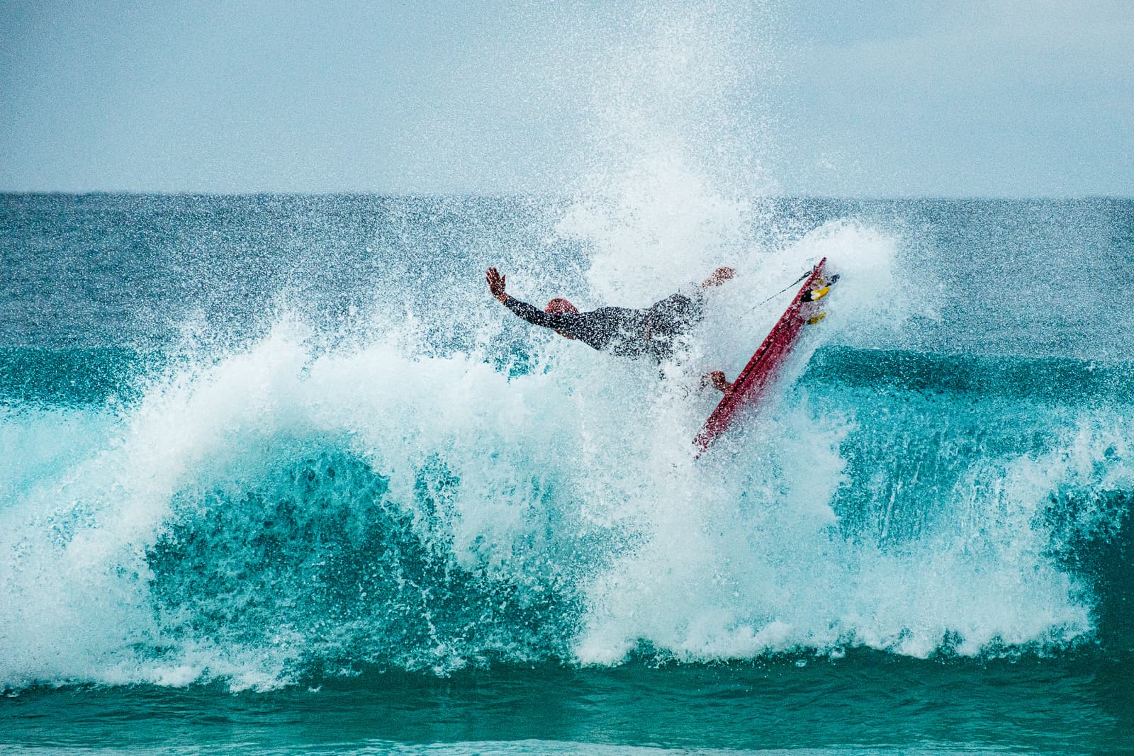 A surfer in a dark wetsuit is mid-air while performing a high-speed maneuver on a wave at Pipeline, their body outstretched with arms wide against a massive plume of sea spray, with a red surfboard visible beneath them.