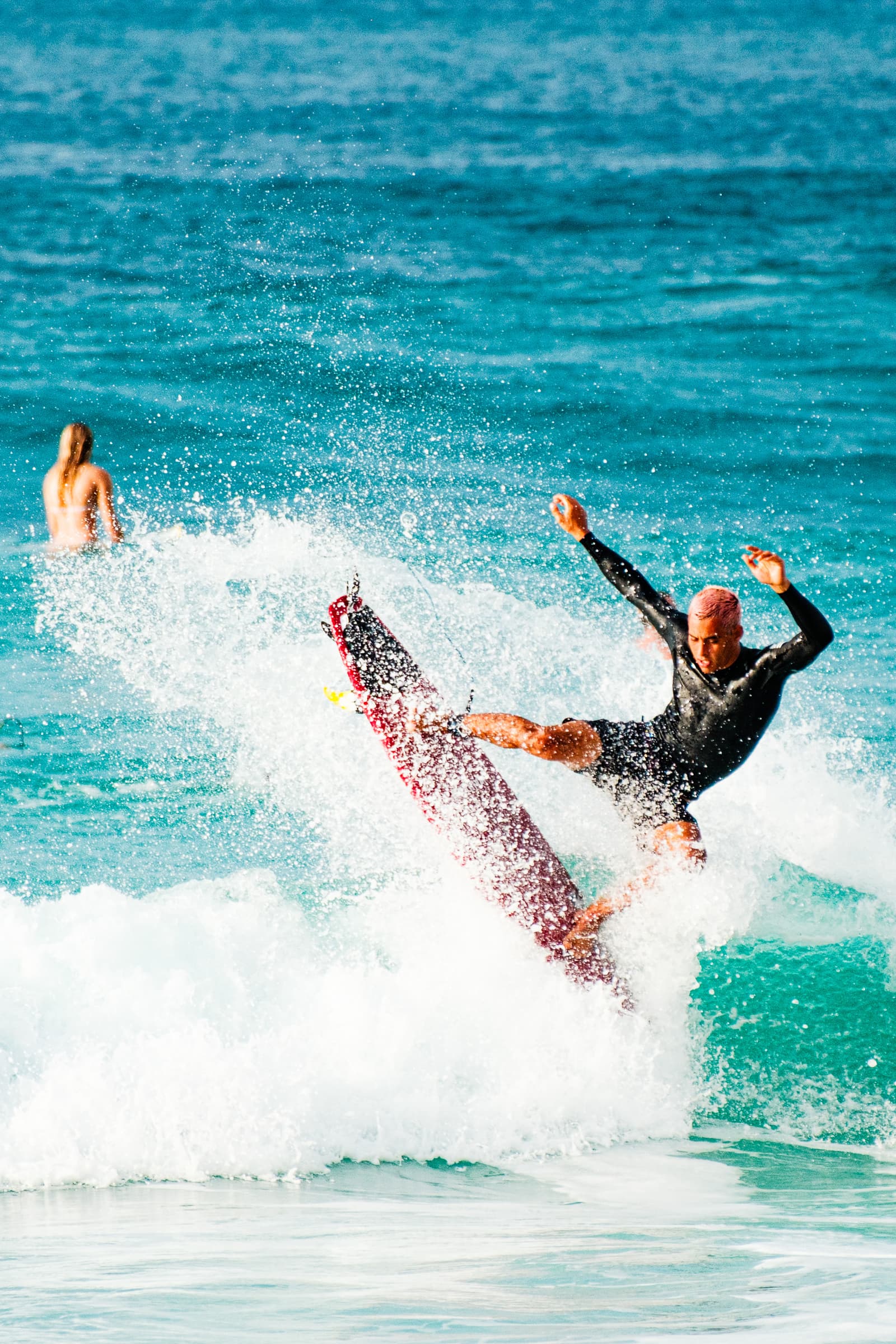 A person with short, light-colored hair and a black wetsuit performs a sharp turn on a red surfboard in an ocean wave. Water spray is ejected behind the board and the surfer. In the background, another person is partially visible in the water.
