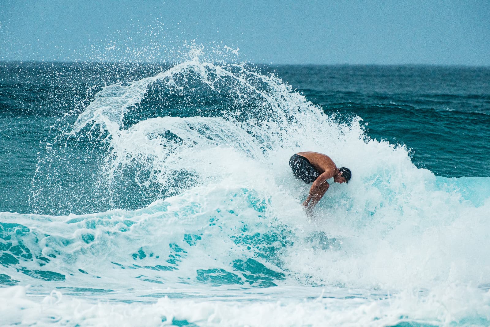 A person surfing a wave on a surfboard, leaning into a sharp turn, with a large spray of white water rising into an arch behind them. The water is deep turquoise and dark blue, contrasted by the bright white foam of the cresting wave.