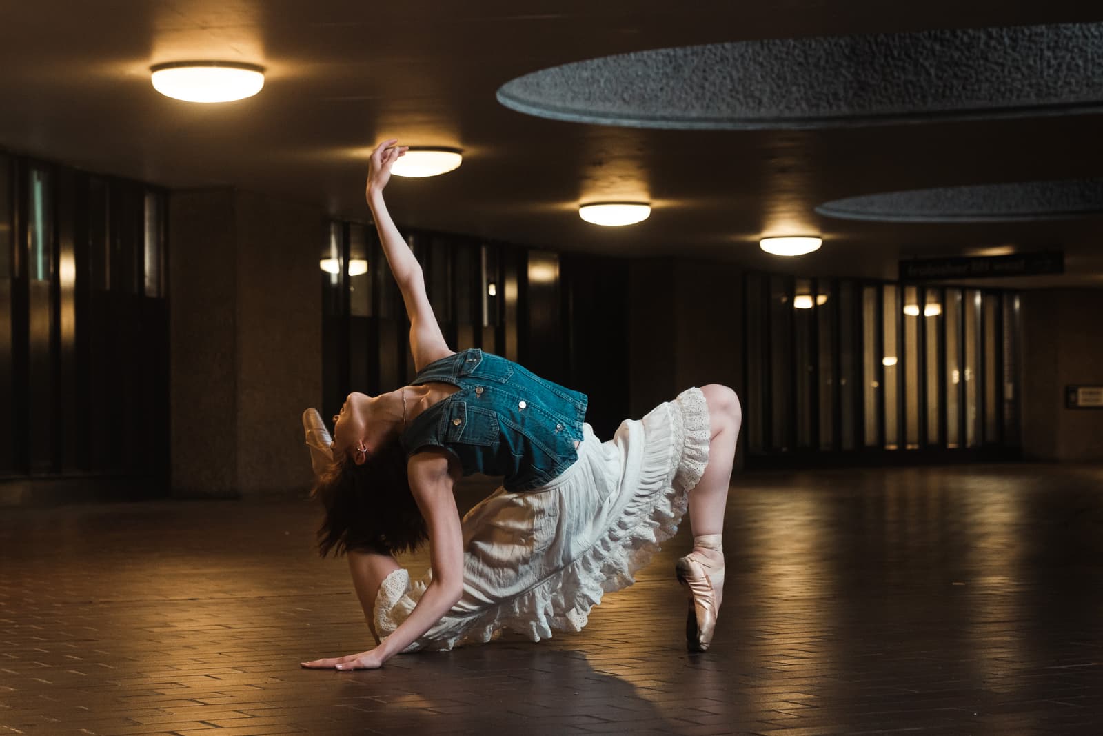 A person in a denim vest and ruffled white skirt performs a ballet floor pose inside a dimly lit concrete parking structure with overhead circular lights. The person is arched backward with one arm raised and one leg tucked towards their back, wearing pointe shoes on a dark tiled floor.