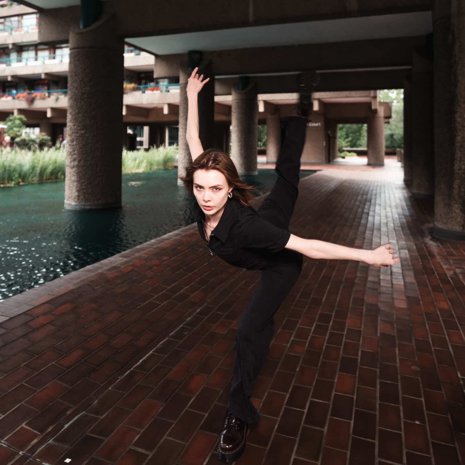 A person stands on one leg in a dance pose with the other leg extended high into the air. They are wearing a black short-sleeved collared shirt, black trousers, and dark platform shoes. The background is a brutalist architecture setting with dark concrete columns, a small pond, and paved ground under a covered walkway.