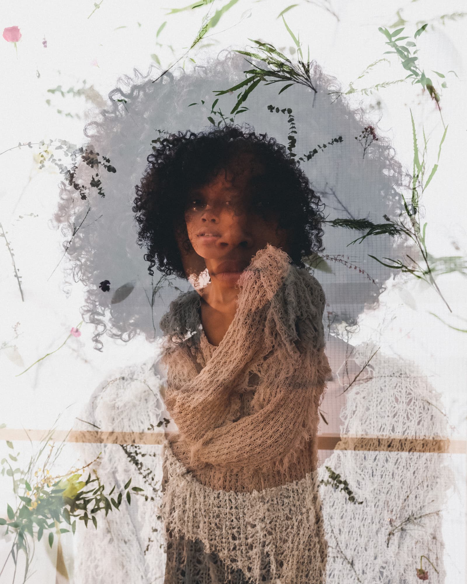 A double-exposure portrait featuring a subject with dark curly hair wearing a textured, loose-knit, distressed beige sweater, layered over a blurred background of white fabric and silhouettes of pressed flowers and foliage.