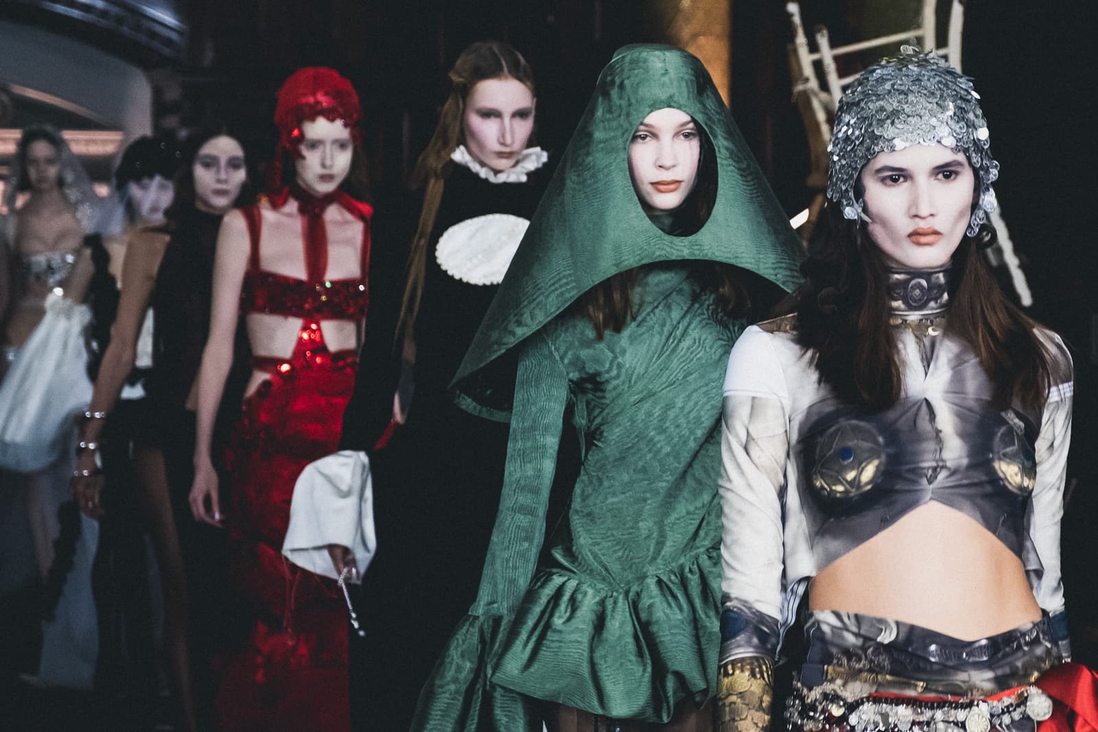 A line of models walking a dimly lit catwalk. In the foreground, a model wears a sequined silver headpiece, a cropped top with a printed design, and elaborate metalwork ornamentation at the neck. Behind her, another wears a structured, pointy green hood over a long-sleeved textured green garment. Further back, models in red and black designs walk in sequence, with various intricate headpieces and textural fabrics visible.