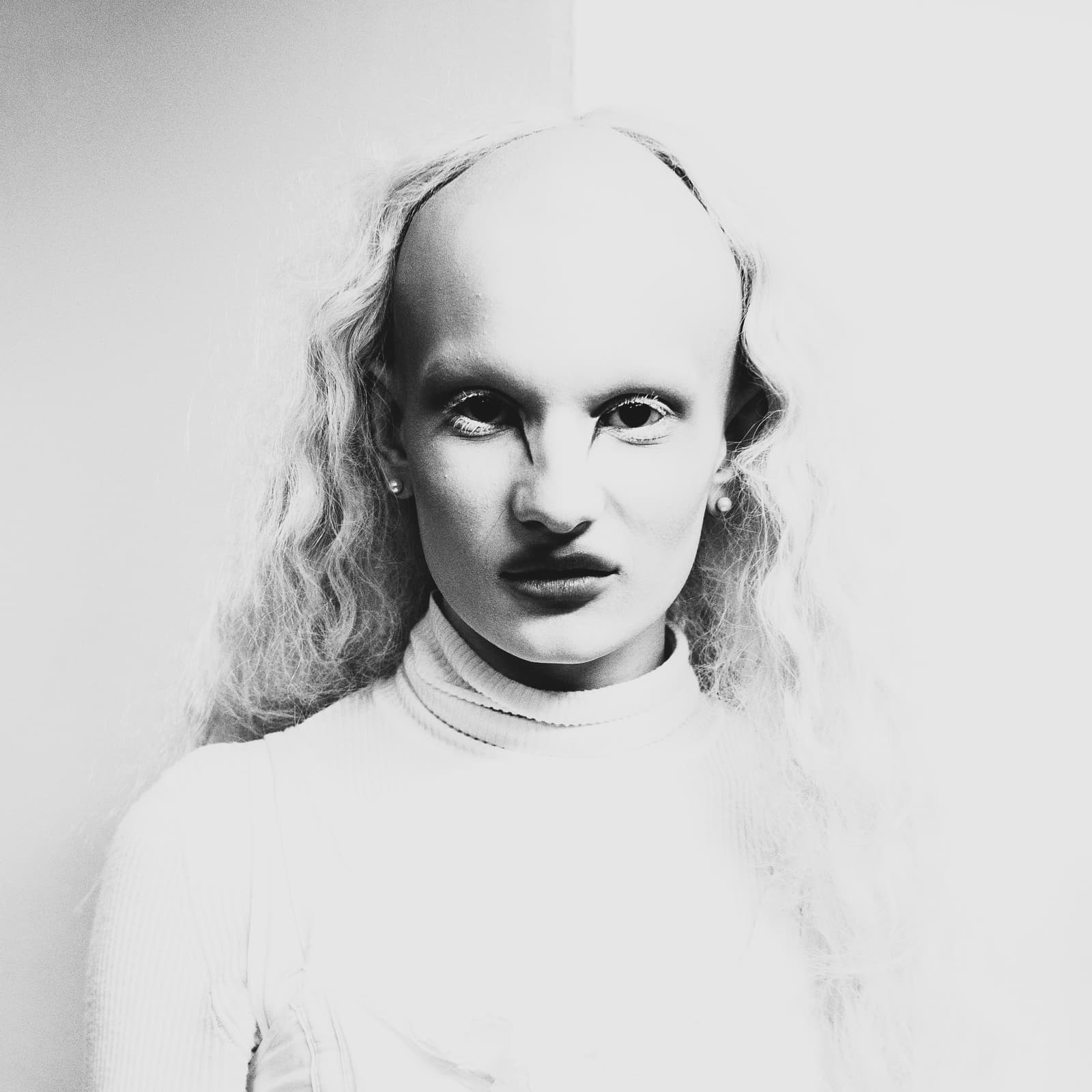 A black-and-white portrait of a person with a shaved head and long, textured blonde hair framing the sides. They have bleached, pale skin, thin eyebrows, and dark, sharp eyeliner applied at the inner corners of their eyes. They are wearing a plain, white, ribbed, high-neck garment against a solid, light-colored background.