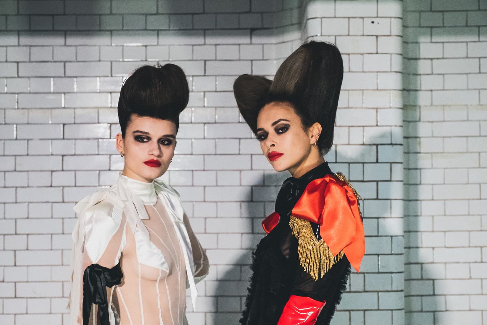 Two individuals posing against a white-tiled wall, both featuring elaborate, voluminous hairstyles and dramatic makeup including dark eyeshadow and bold red lips. The person on the left wears a sheer, light-coloured garment with white ribbon detailing, while the person on the right wears a black top accented with a large red satin bow and gold fringe epaulettes.