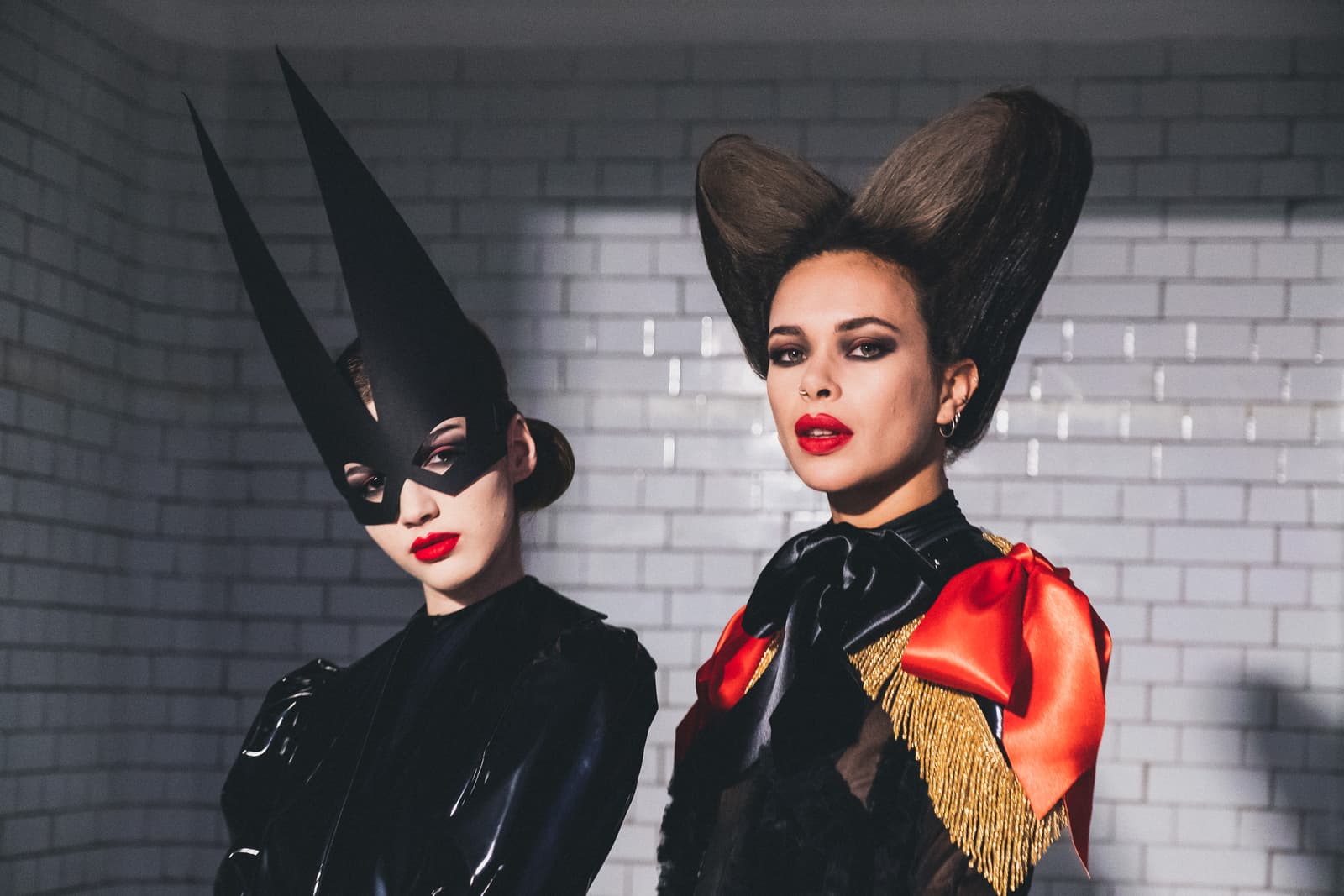 Two people posed against a white tiled wall. The person on the left wears a black latex top and a tall, pointed black mask over the eyes. The person on the right has hair styled into two prominent, rounded peaks and wears a black top with gold fringe detailing and a large red bow. Both have bold red lipstick.