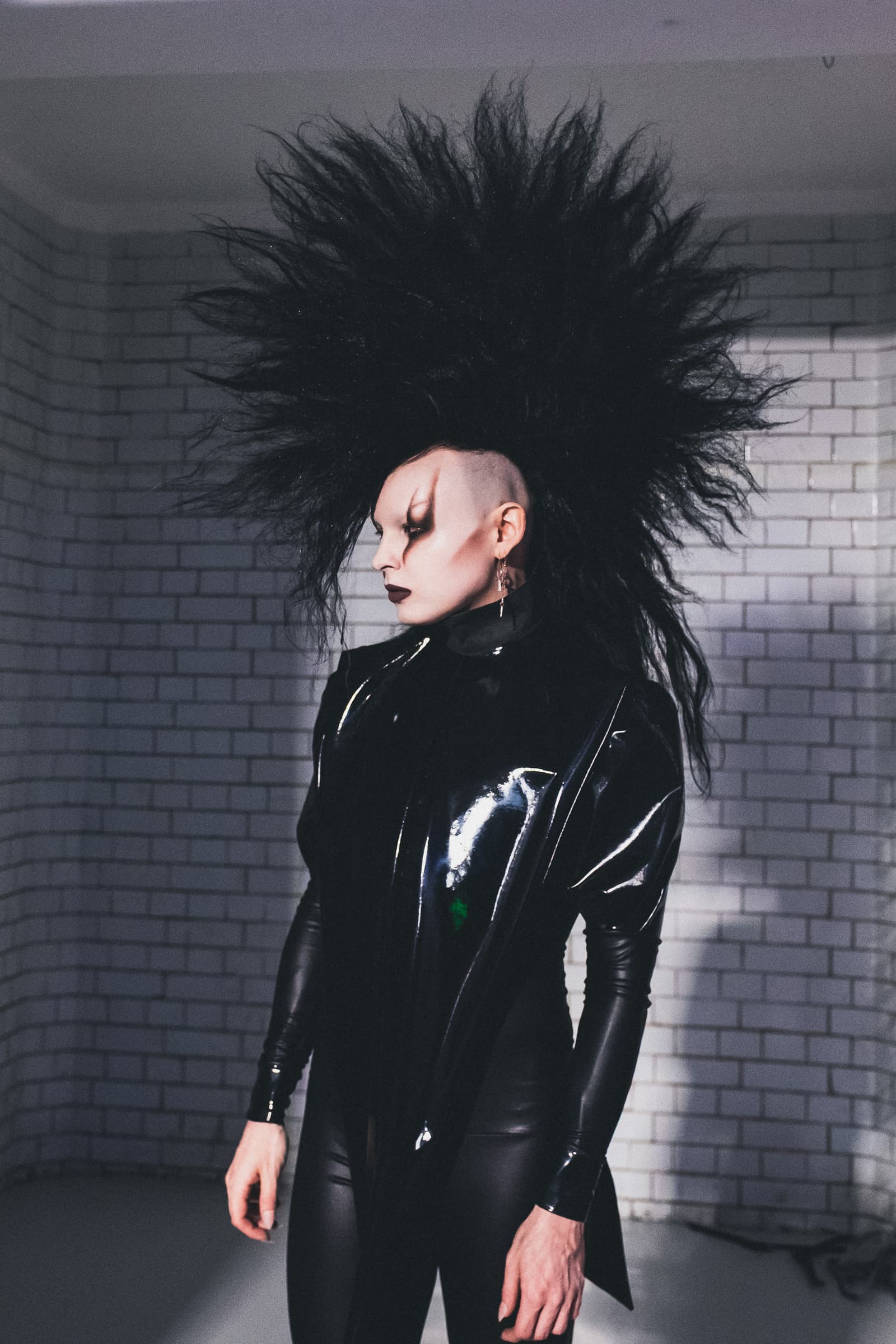 A person with a dramatic, massive black spiked hairstyle stands in profile against a grey brick wall. They are wearing a form-fitting black latex jacket with long sleeves over dark trousers. Their makeup includes heavy dark eye shadow and dark lipstick, and the side of their head is closely shaved.