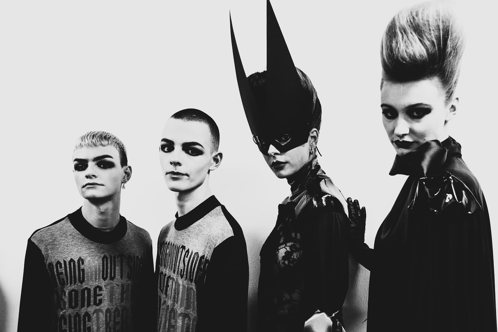 A black-and-white medium shot of four people standing in a line against a plain white background, likely backstage at a fashion show. The two people on the left have short hair and wear grey tops with text. The person second from the right wears a tall, pointed black headpiece or mask with cut-outs over the eyes. The person on the far right has a high, structured beehive hairstyle, dark eyeshadow, and a dark high-collared garment. All four figures are looking toward the camera.