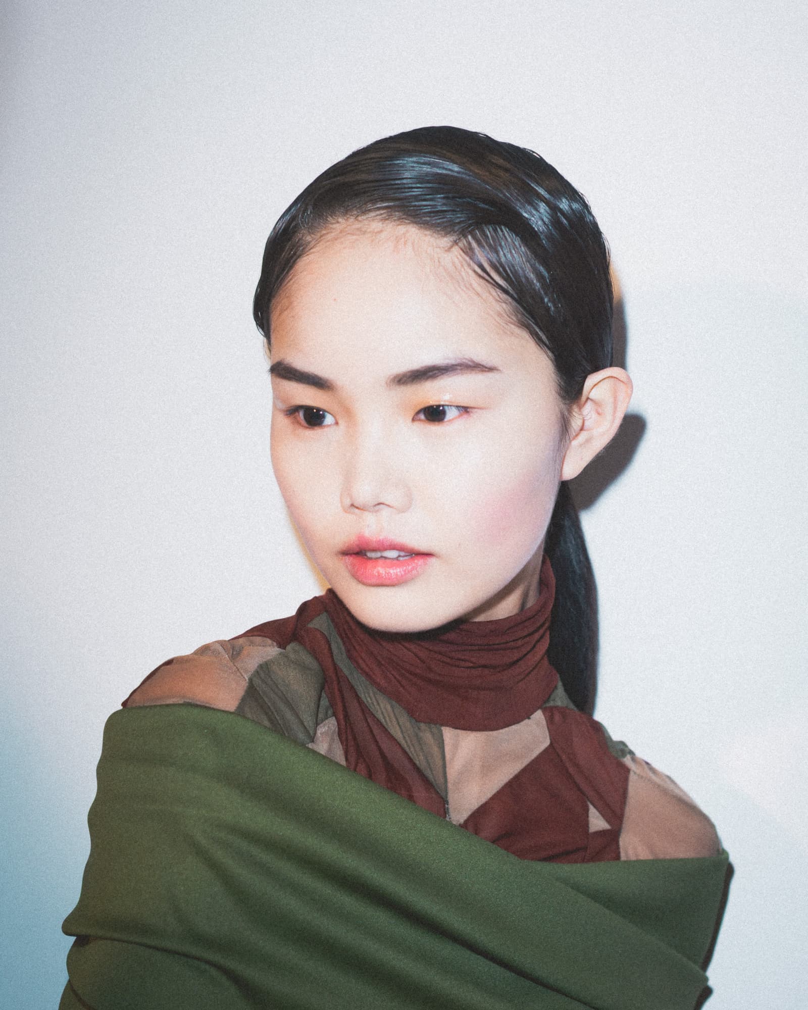 Close-up portrait of a person with dark hair pulled back, wearing an olive green fabric wrapped around the shoulders over a brown and khaki patterned high-neck garment, looking off-camera against a plain light wall under direct studio lighting.