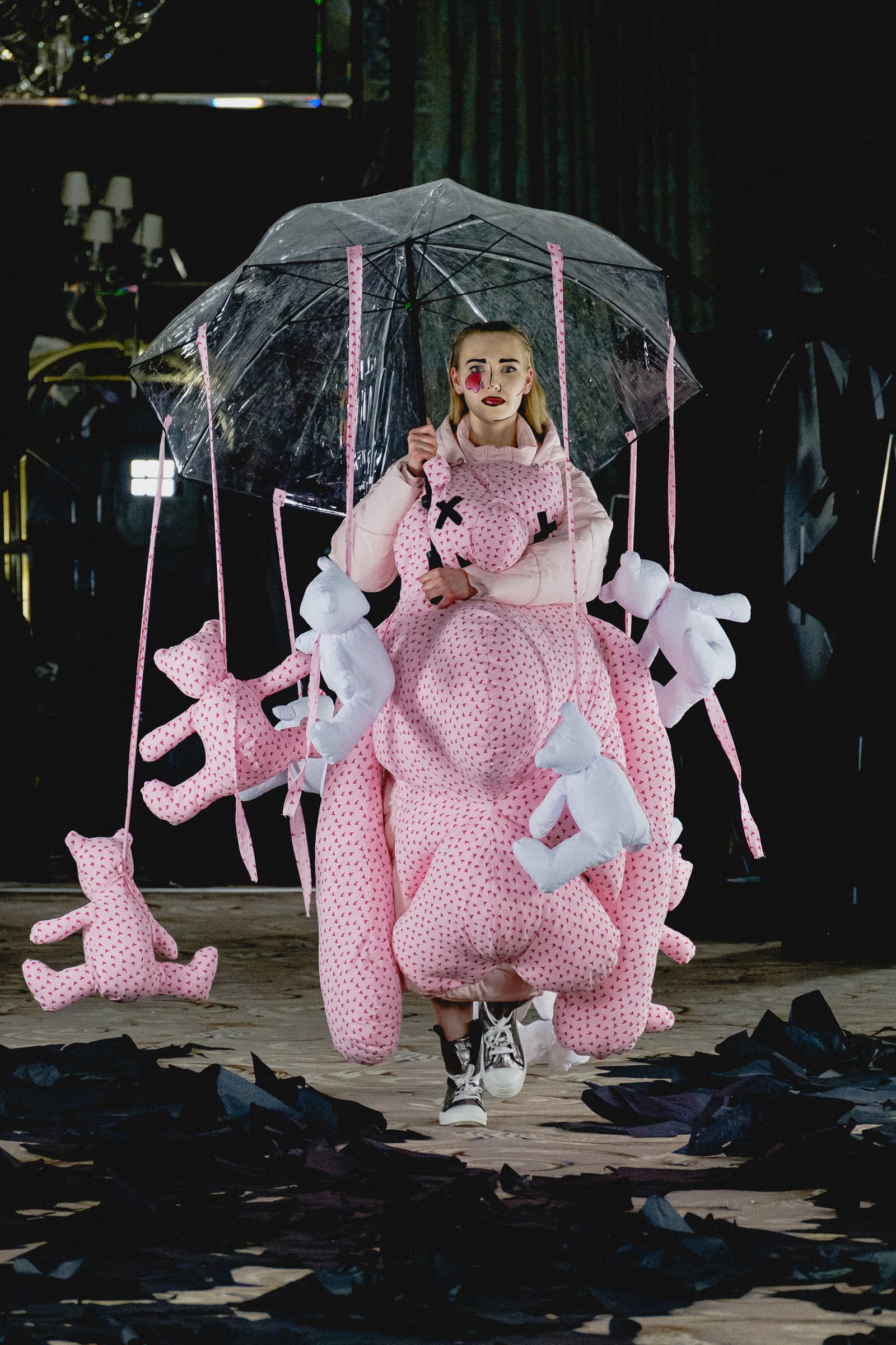 A person walking on a runway, wearing a voluminous, padded pink garment covered in small darker patterns, with several plush teddy bears suspended from a clear umbrella held above their head. The person has a heart-shaped sticker on one cheek and is wearing black and white high-top sneakers, against a dark, industrial-style background with black paper scattered on the floor.