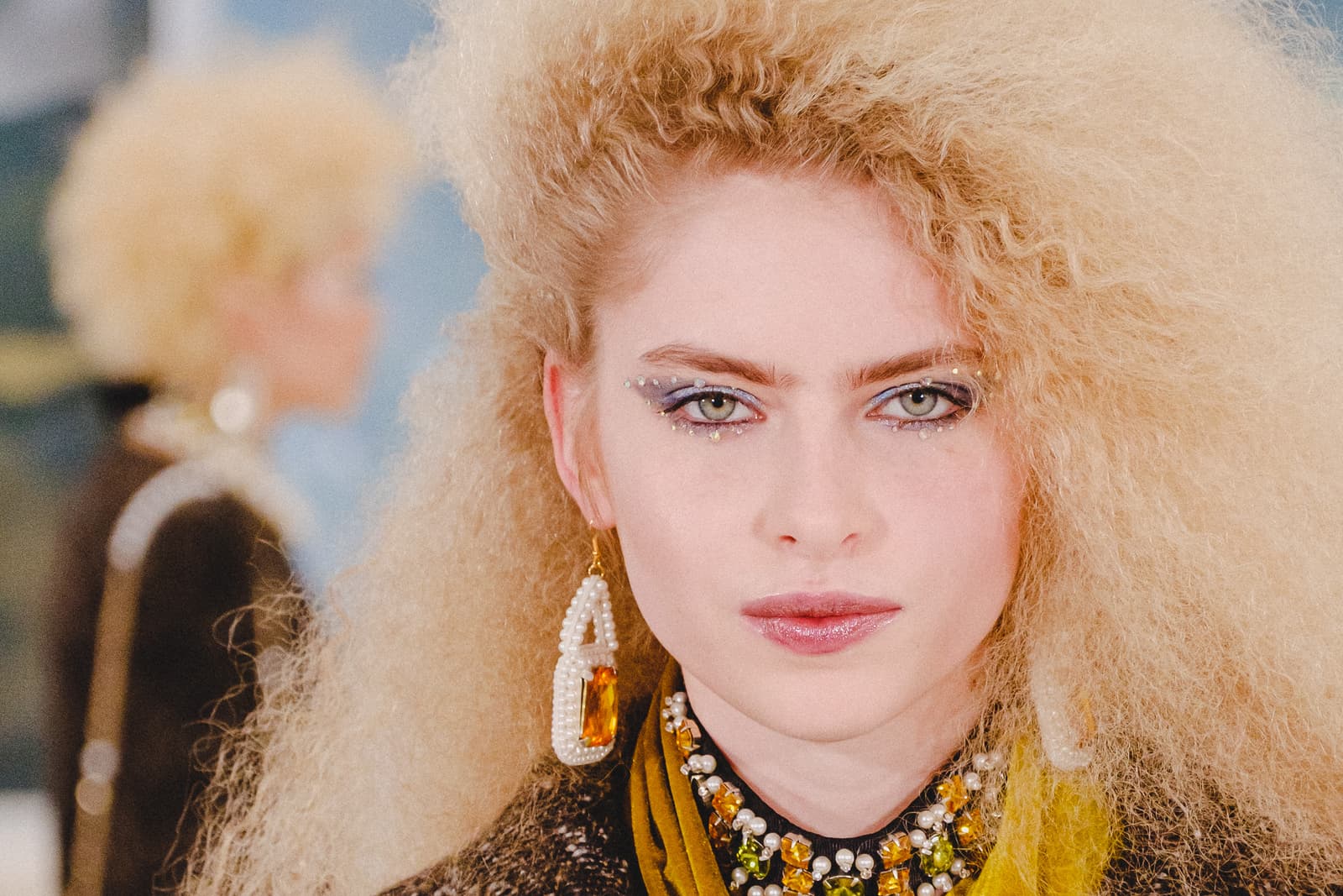 A close-up portrait of a person with voluminous, crimped blonde hair facing the camera. They are wearing ornate pearl and amber jewelry around the neck and a large pearl-encrusted amber earring. Their eye makeup features sharp, winged eyeliner accented with small jewels. Another person with similar hair is partially visible and out of focus in the background.