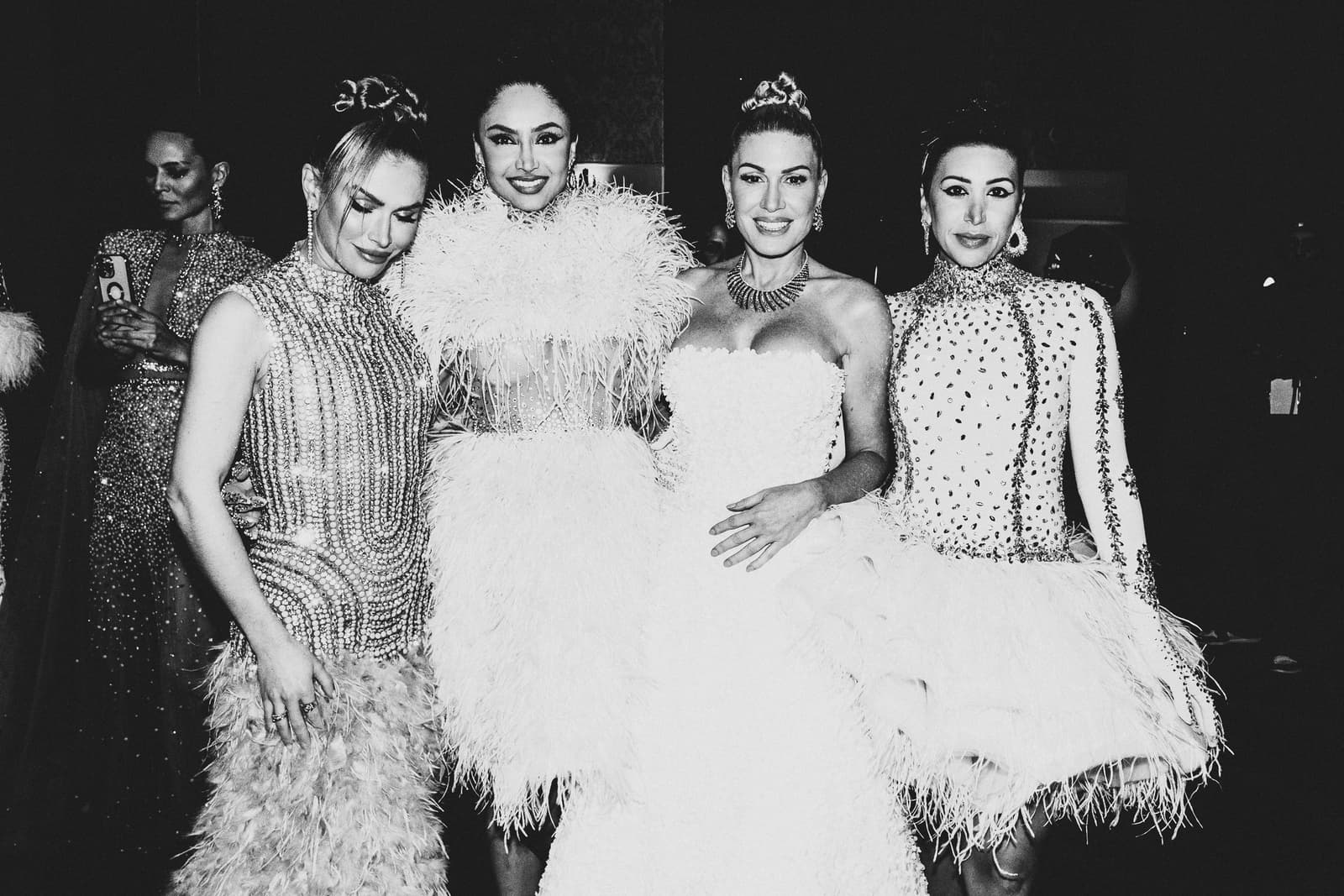A black-and-white medium shot of four people posing together backstage at a fashion show. The people wear elaborately embellished evening dresses, including garments covered in sequins, beads, and white feathers. They are styled with elaborate updos and statement jewelry. In the background, an additional figure is partially visible holding a smartphone.