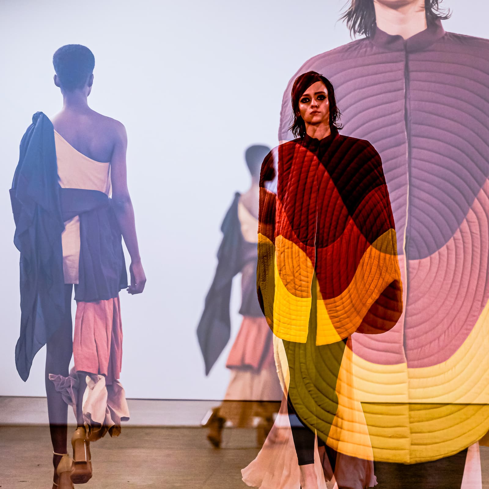 A double-exposure photograph from a fashion runway. In the foreground, a person with dark hair and heavy makeup wears a quilted, collarless jacket featuring concentric circular patterns in shades of yellow, red, and deep purple. Partially overlapping, a person viewed from behind walks down a white runway, wearing a sleeveless, asymmetric top with deep blue fabric draped over one shoulder and light-toned trousers or skirt. The background is a bright, neutral studio space.