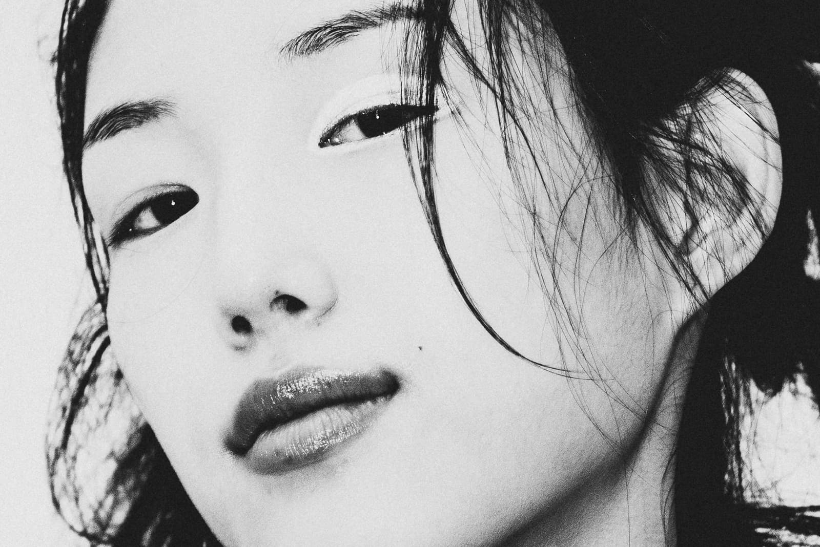 A close-up black and white portrait featuring the face of a person with dark hair, a soft gaze toward the camera, and detailed makeup around the eyes and lips, illuminated by bright, even studio light.