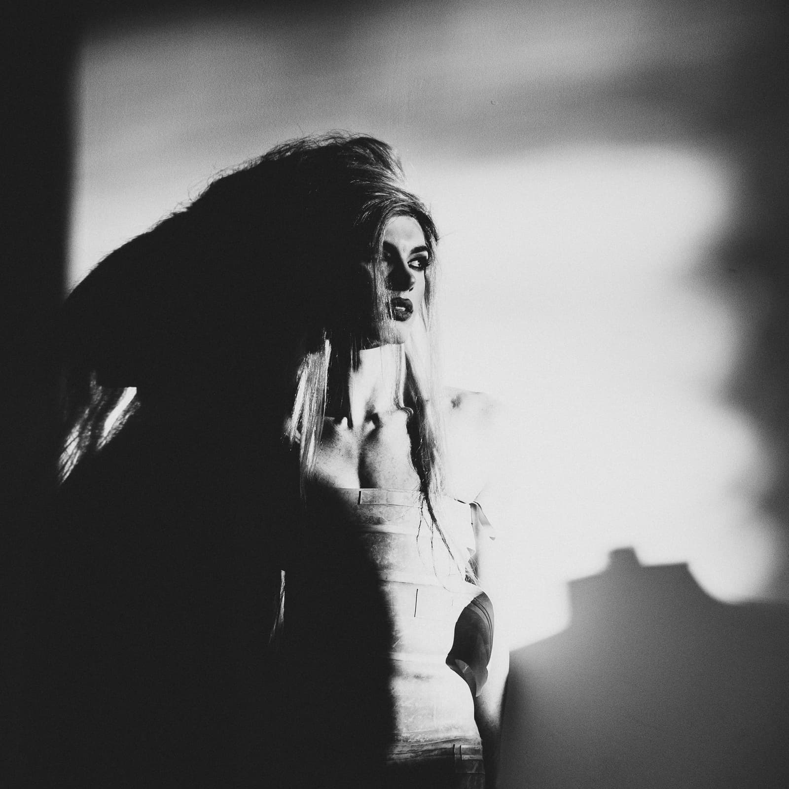 A black-and-white medium close-up of a person with high-volume, teased hair. The individual wears a structured, bodice-like garment with visible panels. Intense side lighting casts a heavy shadow across half of the face and creates a dramatic silhouette on the wall behind. The gaze is directed away from the camera.