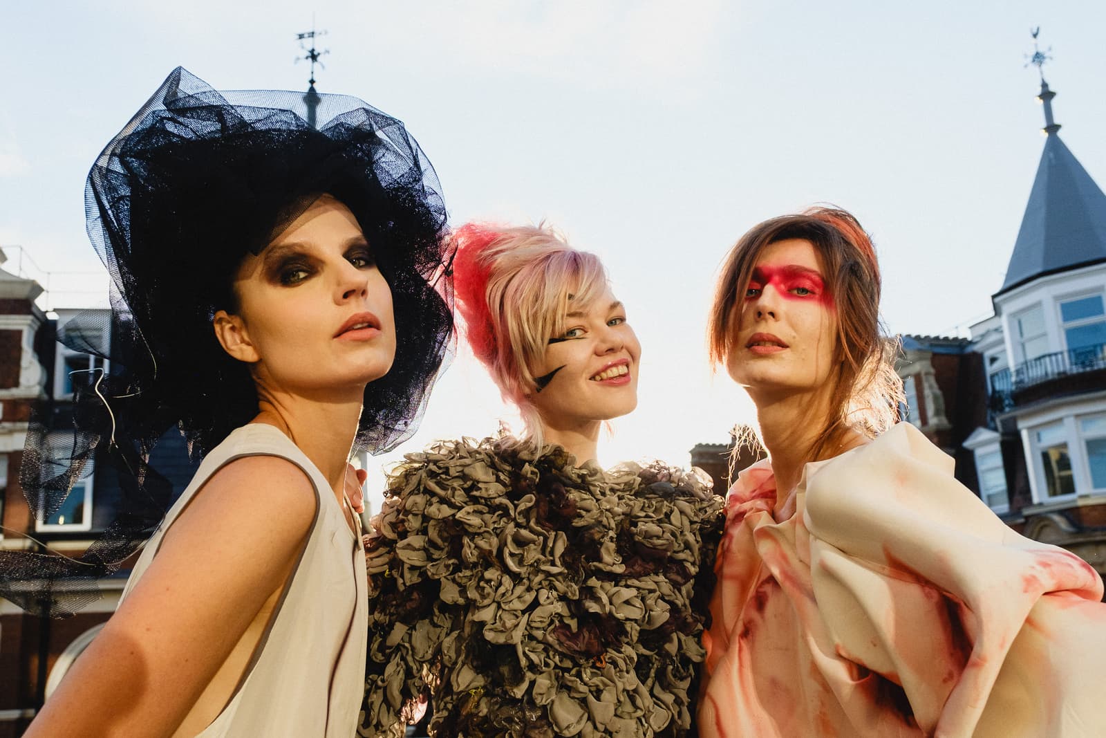 Three individuals positioned shoulder-to-shoulder, shot from a low angle against a clear sky and architectural rooftops. The person on the left wears an oversized black tulle headpiece and dark eye makeup. The person in the center has styled, highlighted hair and a textured, ruffled brown garment. The person on the right features bold pink makeup across their eyes and a pale, draped garment. They are all looking towards the camera.