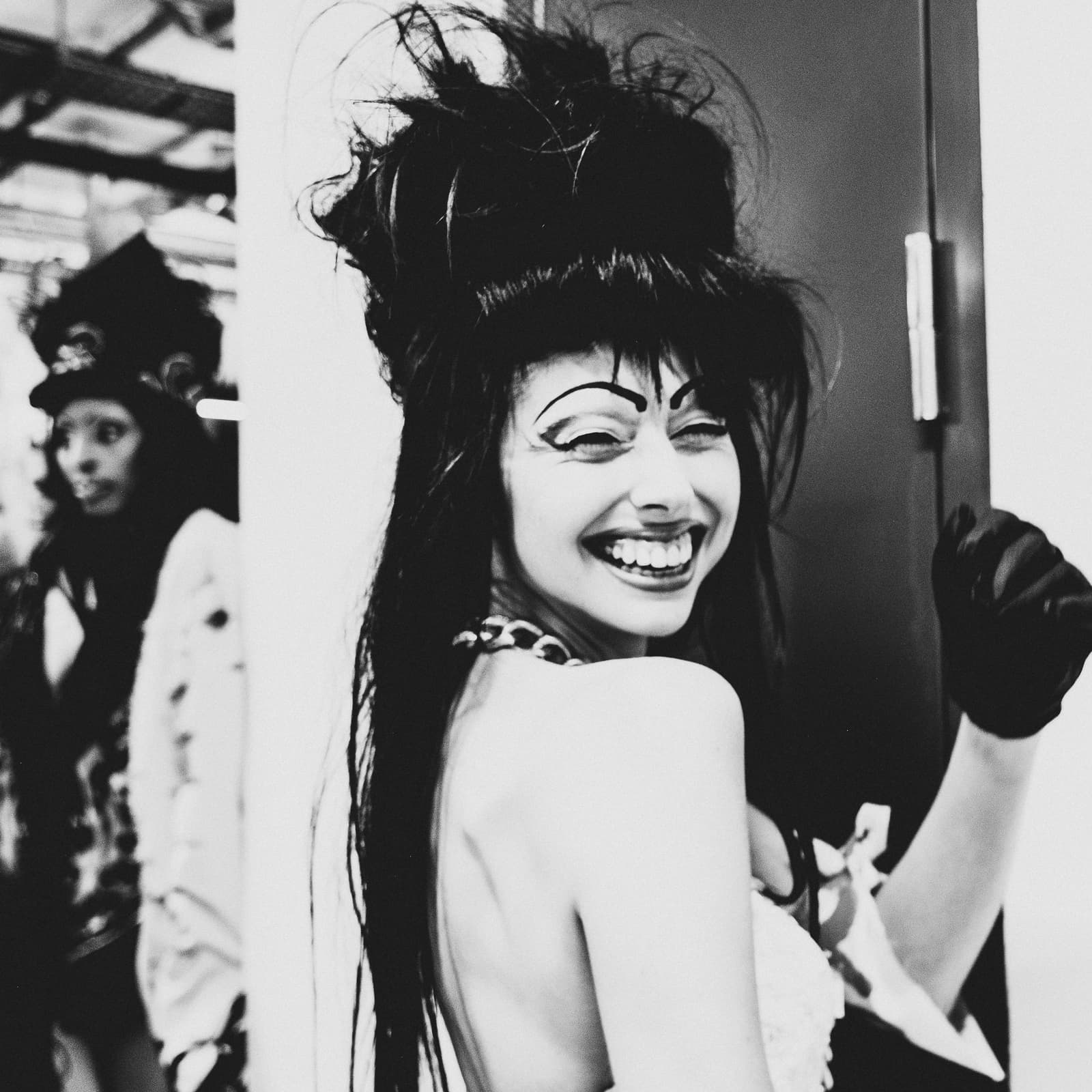 A black-and-white medium shot taken backstage at a fashion show. In the foreground, a smiling person with high-teased dark hair and arched, theatrical makeup looks over their shoulder at the camera, wearing a white sleeveless garment and a black glove on one raised hand. Another person in a dark hat and white coat is visible in the blurred background to the left, standing near a wall.