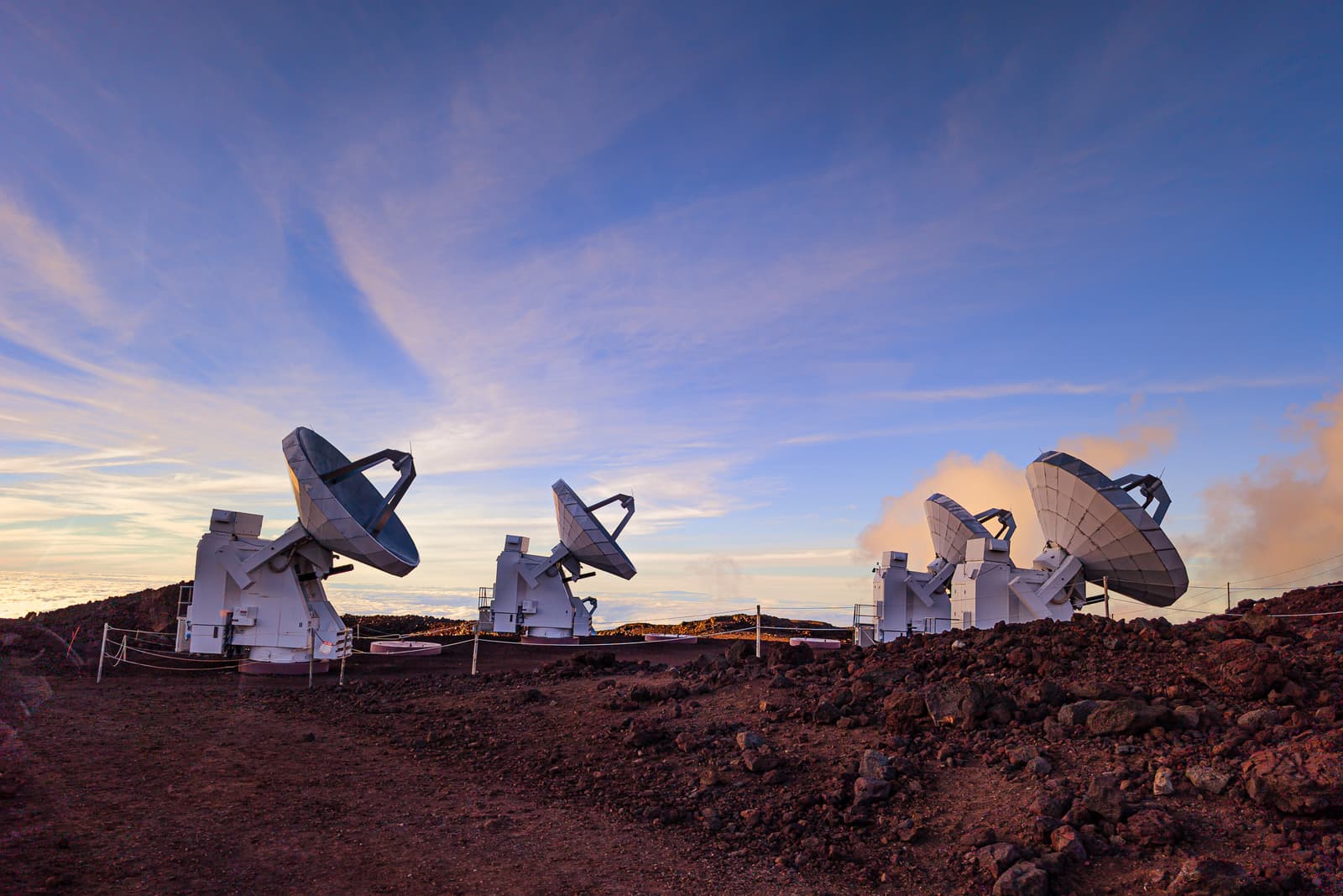 A wide angle view of several large white satellite dish antennas positioned on a dark reddish brown volcanic landscape under a clear blue sky with thin wispy clouds. The scene is lit by golden hour light hitting the clouds and the volcanic terrain, with a small rope barrier in the foreground and a sea of clouds visible in the distance behind the equipment.