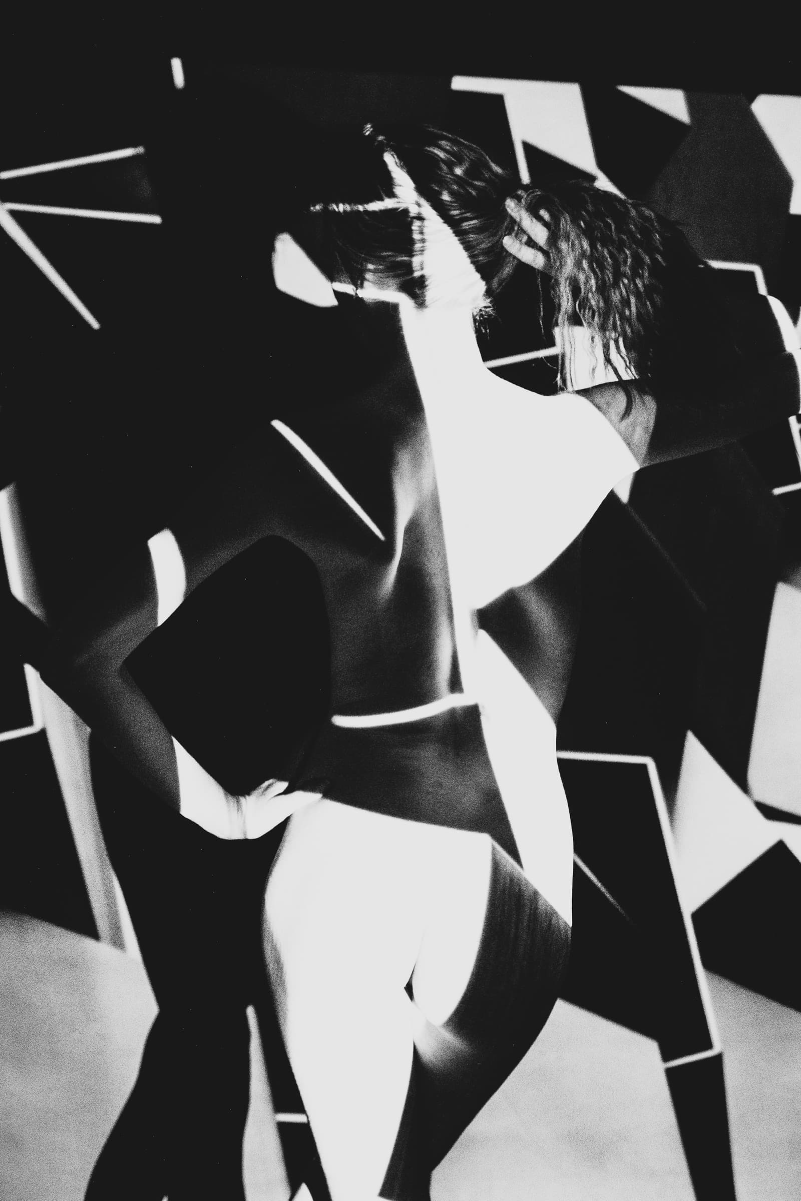 A black and white high-contrast studio photograph featuring a person partially obscured by geometric, solarised light projections. Sharp angles, blocks of black, and thin white lines overlay the figure, who has their hands raised near their head. The subject's form is fragmented by the light, creating an interplay of solid shapes and organic curves.
