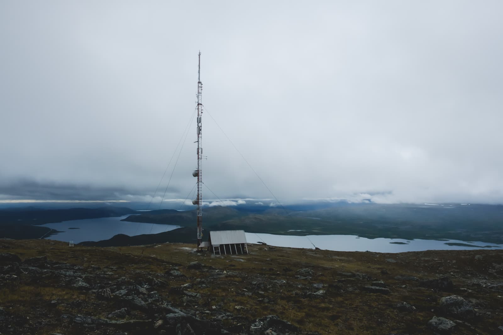 A tall, slender telecommunications tower stands on a barren, rocky mountain ridge under a heavy, overcast sky. Below the tower is a small, shed-like wooden structure. In the background, a vast, dark blue lake stretches across a valley, surrounded by rolling hills and distant, low mountains fading into mist.